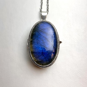 May include: An oval locket pendant featuring a large, polished labradorite gemstone with deep blue and golden iridescence. The stone is set in a silver-toned metal frame with a beaded edge, suspended from a matching silver-toned chain link necklace.