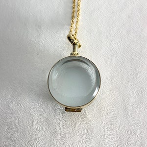 May include: A gold-colored magnifying glass pendant necklace with a clear, round lens. The pendant has a hinged bottom and hangs from a delicate gold chain. The background is a textured white surface.