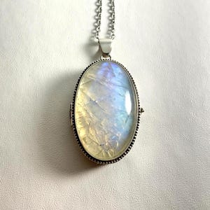 May include: An oval locket pendant featuring a large, iridescent moonstone cabochon with visible internal fractures and blue light reflections, set in a silver-toned metal frame with a beaded edge, suspended from a matching silver-toned chain.