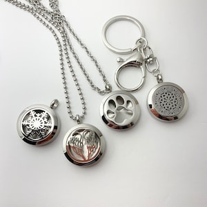 May include: Four silver essential oil diffuser necklaces with different designs. One necklace has a snowflake design, another has a paw print, another has a wing design, and the last one has a circular design. The necklaces are on a white background.