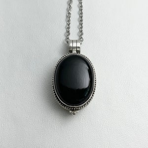 May include: A silver-toned necklace featuring a large, oval, black gemstone pendant. The pendant is set in a detailed bezel with a beaded edge and hangs from a delicate chain. The gemstone has a glossy, reflective surface.