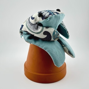 May include: A handmade frog-shaped plush toy with a patterned head and light blue body. The frog has black button eyes and is perched on a small terracotta pot. The fabric features a blue, white, and navy design.