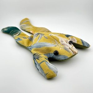 May include: A stuffed animal shaped like a frog, crafted from mustard yellow fabric featuring a botanical print of grey and white leaves. The toy has two black circular button eyes and a solid olive green fabric on its underside and limbs.