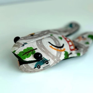 May include: A grey and white fabric toy shaped like a frog with black button eyes and a road and car pattern.