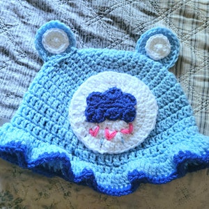May include: A light blue crochet bucket hat featuring two small ears with white centers. The front displays a large white circular patch with a dark blue cloud and three small pink stitched raindrops hanging below the cloud. The brim has a dark blue edge.