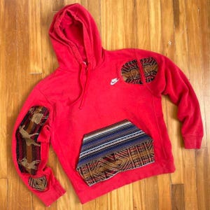 Op de afbeelding: Rode hoodie met een Nike-logo op de linkerborst. De hoodie heeft een grote zak aan de voorkant met een kleurrijke, gepatenteerde voering. De mouwen hebben ook kleurrijke, gepatenteerde stofpatches.
