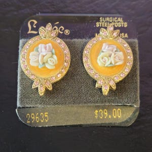 May include: A pair of gold-toned stud earrings. Each earring features a round, orange-colored center with a cluster of small, white and pink roses. The earrings are surrounded by pink rhinestones and have a decorative gold-toned design at the top and bottom.