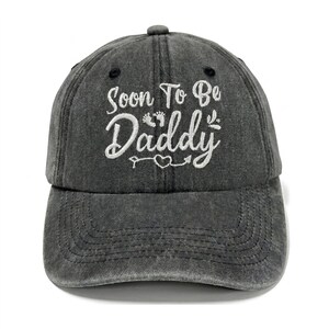 Puede incluir: Una gorra de béisbol gris carbón con acabado desgastado que presenta un bordado blanco con el texto Soon To Be Daddy, acompañado de pequeños iconos de huellas de bebé y un diseño de corazón con una flecha en el panel frontal.