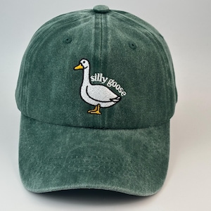 May include: A dark green baseball cap with a washed, distressed texture. The front features an embroidered graphic of a white goose with an orange beak and feet, accompanied by the white embroidered text "silly goose" in a curved font.