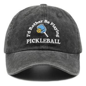 May include: A charcoal grey baseball cap with an embroidered design featuring a blue pickleball paddle and a yellow ball. The text "I'd Rather Be Playing PICKLEBALL" is stitched in white thread in an arched formation above the paddle graphic.