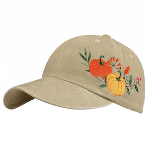 May include: A tan baseball cap featuring an embroidered design on the side. The embroidery depicts two pumpkins, one orange and one yellow, surrounded by green leaves, small red berries, and a single orange leaf, all stitched in vibrant thread.