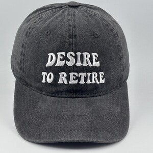Op de afbeelding: Een antracietgrijze honkbalpet met een gewassen, verweerde textuur. De voorkant is voorzien van wit geborduurde tekst met de woorden "DESIRE TO RETIRE" in een vetgedrukt, licht golvend lettertype. De pet heeft een gestructureerde kroon.