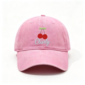 Puede incluir: Una gorra de béisbol de color rosa claro con un diseño bordado de dos cerezas rojas con hojas verdes. Debajo de las cerezas, la palabra Elarly está cosida en una fuente cursiva blanca. La gorra tiene una corona estructurada y visera curva.