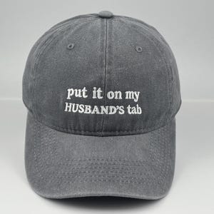 Könnte beinhalten: Eine anthrazitfarbene Baseballkappe mit weißem, gesticktem Text auf der Vorderseite, der "put it on my HUSBAND'S tab" lautet. Die Kappe hat eine strukturierte Krone, einen gebogenen Schirm und genähte Ösen zur Belüftung an den oberen Paneelen.