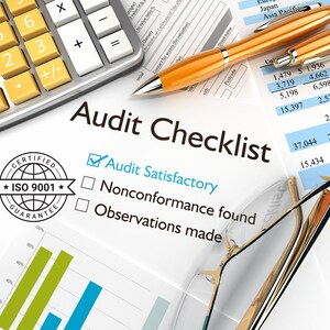 Op de afbeelding: Een witte checklist met de tekst "Audit Checklist" en drie selectievakjes. Het eerste selectievakje is aangevinkt en heeft het label "Audit Satisfactory". Het tweede selectievakje is niet aangevinkt en heeft het label "Nonconformance found". Het derde selectievakje is niet aangevinkt en heeft het label "Observations made". Er is ook een rekenmachine, een pen en een bril op de afbeelding.