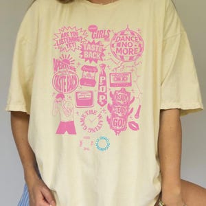May include: Cream-colored t-shirt with a pink graphic design. The design includes phrases like "ARE YOU LISTENING?" and "DANCE NO MORE," along with other illustrations. The shirt has a relaxed fit.