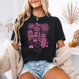 May include: Black t-shirt with a pink graphic design featuring text and illustrations. The design includes phrases like "Girls Taste Back" and "Dance No More." The shirt is paired with denim shorts and a cream-colored cardigan.