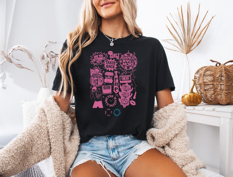 May include: Black t-shirt with a pink graphic design featuring text and illustrations. The design includes phrases like "Girls Taste Back" and "Dance No More." The shirt is paired with denim shorts and a cream-colored cardigan.