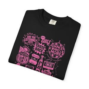 May include: Black t-shirt with a pink graphic design featuring text and illustrations. The design includes phrases like "ARE YOU LISTENING?" and "DANCE NO MORE," along with other words and images. The shirt is folded.