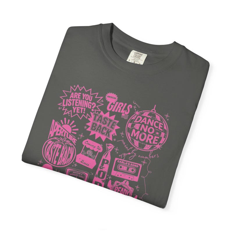 May include: Dark grey t-shirt with a pink graphic design. The design includes text such as "ARE YOU LISTENING?" and "AMERICAN GIRLS", along with other phrases and images. The shirt is folded.