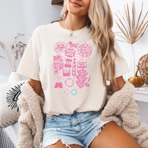 May include: Cream-colored t-shirt with a pink graphic design featuring text and illustrations. The design includes phrases like "Girls," "Dance No More," and "Are You Listening Yet?" The shirt is paired with denim shorts.