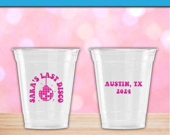May include: Two clear plastic disposable cups. The left cup features a pink graphic of a disco ball with the text SARA'S LAST DISCO. The right cup displays the pink text AUSTIN, TX 2024. Both cups are transparent with a smooth, tapered design.