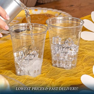 May include: Two clear plastic cups filled with ice cubes, each featuring black printed text that reads The Morans EST. 2027. One cup is being filled with liquid from a silver container, showcasing the transparent design and the custom lettering.