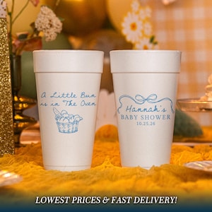 May include: Two white foam cups featuring blue text and graphics. The left cup displays the text A Little Bun is in The Oven with a basket of bread. The right cup shows a blue ribbon graphic, the text Hannah's BABY SHOWER, and 10.25.26.