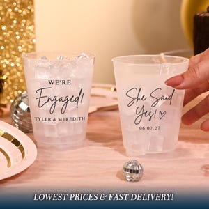 May include: Two frosted plastic cups filled with ice cubes. The left cup features the black text "WE'RE Engaged!" and "TYLER & MEREDITH". The right cup features the black text "She Said Yes!" with a heart symbol and the date "06.07.27".
