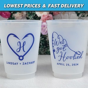 May include: Two white plastic cups with blue text and designs. One cup says "We got Hooked" with a fishing rod and hook design and the date "April 25, 2024". The other cup has a heart with a fishing hook design and the letter "H" inside the heart. The text below the heart says "Lindsay + Zachary".