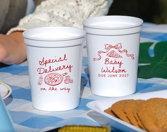 May include: Two white plastic cups featuring red printed graphics. The left cup displays the text Special Delivery on the way with a baby bottle, pizza, and blocks. The right cup shows a bow, a pizza slice, and the text Baby Wilson DUE JUNE 2027.