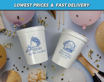 May include: Two white plastic cups with blue designs. One cup says "Fresh off the market" with a blue illustration of a basket of flowers. The other cup says "Amanda's Bridal Shower 05.24.25" with a blue illustration of a basket of flowers.