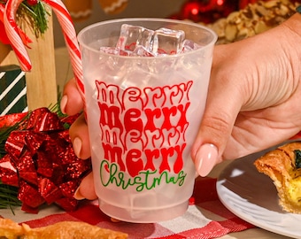 May include: A clear plastic cup filled with ice cubes, featuring the red text "merry merry merry merry" stacked vertically and the green text "Christmas" underneath, with a candy cane visible on the left side of the cup.