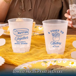 May include: Two clear plastic cups with blue text. One says "Butter Together Mallory's Bridal Shower" and the other says "A Toast To Mallory 05.15.27". The cups are on a yellow tablecloth with a floral plate.