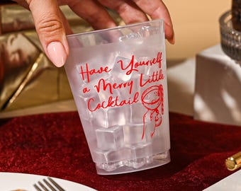 May include: A clear plastic tumbler filled with ice cubes, featuring red cursive text that reads Have Yourself a Merry Little Cocktail and a small red line drawing of a cocktail glass with a garnish, held in a hand with light pink nail polish.