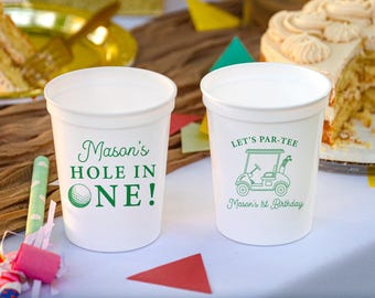 May include: Two white plastic cups featuring green text and graphics. One cup displays "Mason's HOLE IN ONE!" with a golf ball icon. The other cup shows a golf cart graphic with the text "LET'S PAR-TEE" and "Mason's 1st Birthday".