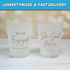 May include: Two white plastic cups with gold lettering. One cup says "We're Engaged!" with the names "Eric & Carolina" below. The other cup says "She Said Yes!" with a heart and the date "10.22.24" below.