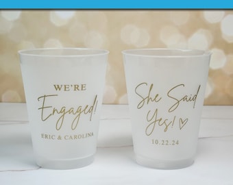 May include: Two white plastic cups with gold lettering. One cup says "We're Engaged!" with the names "Eric & Carolina" below. The other cup says "She Said Yes!" with a heart and the date "10.22.24" below.