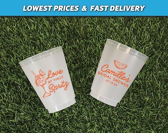 May include: Two frosted plastic cups with orange text and graphics. One cup says "Love at First Spritz" with a cocktail illustration. The other says "Camille's Bridal Shower 07.19.25" with a citrus slice. The cups are on green grass.