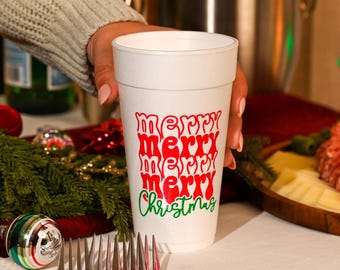 May include: A white foam cup featuring the repeating red text "merry" four times in a wavy, retro-inspired font, followed by the word "Christmas" in a green cursive script. The cup has a smooth, tapered cylindrical shape and a wide rim.