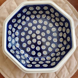 May include: An octagonal ceramic bowl featuring a deep blue interior decorated with a repeating pattern of small white, green, and red concentric circles. The bowl has a thick, smooth white rim and a glossy finish throughout the entire surface.