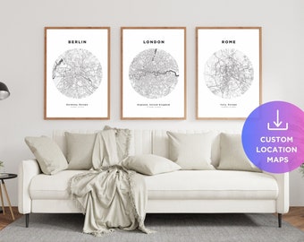May include: Three framed prints displaying city maps of Berlin, London, and Rome. Each map is a black and white circular design with the city name above and the country below. The prints are above a white sofa with beige cushions and a throw.