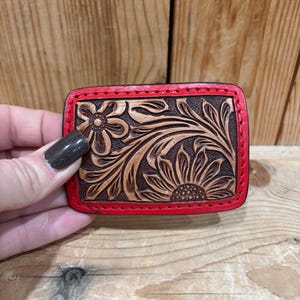 K&ouml;nnte beinhalten: Rechteckige, braune Lederg&uuml;rtelschnalle mit floralem Design. Das Design zeigt eine Sonnenblume und andere florale Elemente. Die Schnalle hat einen roten Rand und wird vor einem Holzgrund gehalten.
