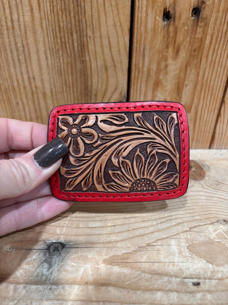 K&ouml;nnte beinhalten: Rechteckige, braune Lederg&uuml;rtelschnalle mit floralem Design. Das Design zeigt eine Sonnenblume und andere florale Elemente. Die Schnalle hat einen roten Rand und wird vor einem Holzgrund gehalten.