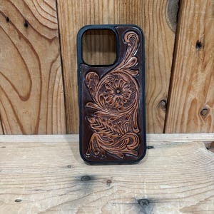 May include: A dark brown leather phone case featuring an intricate, hand-tooled floral and scrollwork pattern. The case has a black protective frame and a rectangular cutout at the top for a camera lens, showcasing detailed craftsmanship.