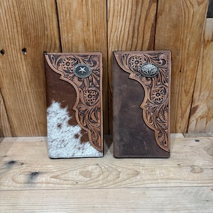 May include: Two rectangular leather wallets featuring intricate floral tooling along the top edges. The left wallet has a brown and white cowhide panel, while the right wallet is solid brown leather. Both include a silver-toned metal concho accent.