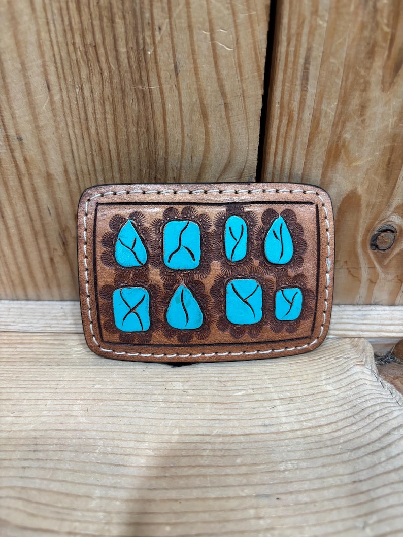 K&ouml;nnte beinhalten: Rechteckige, braune Lederg&uuml;rtelschnalle mit t&uuml;rkisfarbenen Steineinlagen. Die Schnalle hat ein dekoratives Blumenmuster um jeden Stein und wei&szlig;e N&auml;hte am Umfang. Der Hintergrund ist aus Holz.