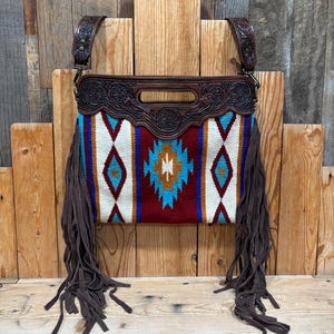 Peut inclure: Sac à bandoulière en cuir marron avec un devant en tissu tissé à motifs géométriques rouges, blancs et turquoise. Le sac a de longues franges marron et des détails en cuir gaufré décoratifs.
