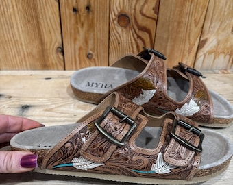 May include: Brown leather sandals with a detailed embossed design featuring feathers and floral patterns. The sandals have two adjustable straps with metal buckles and a cork footbed. The brand name "MYRA" is visible on the footbed.