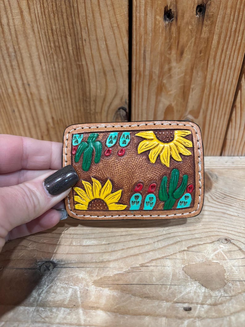 K&ouml;nnte beinhalten: Ein rechteckiger, brauner Lederaufn&auml;her mit handgefertigtem Design mit gelben Sonnenblumen, gr&uuml;nen Kakteen und roten und t&uuml;rkisfarbenen Akzenten. Der Aufn&auml;her wird von einer Hand vor einem Holzgrund gehalten.